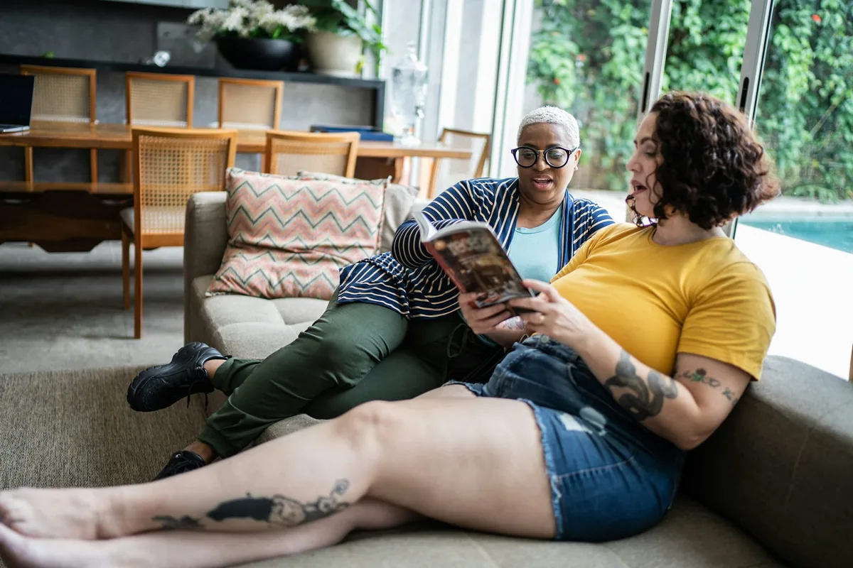 Two women relaxing together on a sofa, one reading a magazine aloud